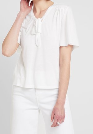 Biały top z krótkim rękawem, okrągłym dekoltem i ozdobą w formie wiązanej kokardy. Tkanina jest lekka i ma gładką teksturę.