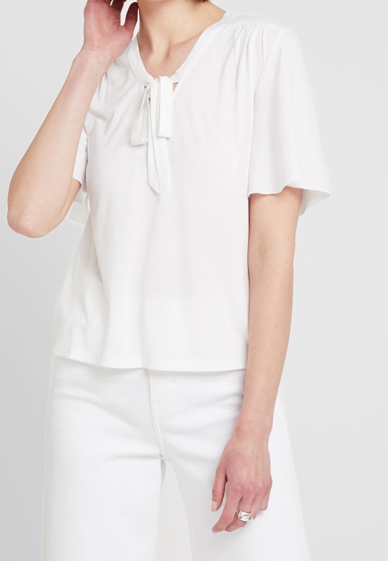 Haut blanc à manches courtes avec un col rond et un détail de nœud papillon. Le tissu est léger et a une texture lisse.