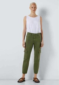 Γυναίκα στέκεται φορώντας μια λευκή αμάνικη μπλούζα, ελαιόχρωμα cargo παντελόνια και καφέ σανδάλια με στρας σε μπροστά φόντο.