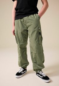 Pantaloni cargo leggeri di colore verde oliva con molte tasche e polsini elastici, abbinati a sneaker nere e bianche, su uno sfondo neutro.