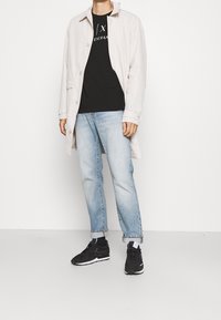 Hellbeige Trenchcoat über einem schwarzen T-Shirt, kombiniert mit hellblauen Jeans und schwarzen Turnschuhen. Der Mantel hat einen Kragen und Knöpfe.