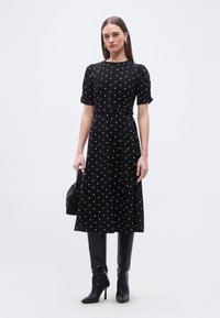 Γυναίκα που φοράει μαύρο midi φόρεμα με λευκές πουά, μανίκια με φουσκωτό στυλ, ζώνη, μαύρες μπότες έως το γόνατο και κρατάει μια μαύρη τσάντα.