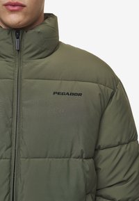 Jachetă puf verde oliv cu guler înalt, având închidere cu fermoar și logo-ul negru „PEGADOR” pe piept. Textură netedă, captusită.