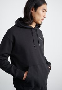 Haut à capuche noir en tissu doux, avec poche kangourou, cordon de serrage ajustable et un petit logo blanc sur la poitrine.