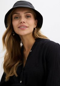 Donna con lunghi capelli mossi, che indossa orecchini a cerchio, un cappello a secchiello nero e una camicia nera con bottoni decorata con ricami floreali, che sorride dolcemente.