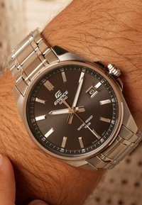 Silver wristwatch with a dark brown face, stainless steel bracelet, and luminous hour markers. Features a date display and water resistance up to 100m.