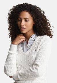 Biely pulóver so vzorom pletenia káblovým vzorom s výstrihom do V, nosený cez modrú pruhovanú košeľu. Má rebrované manžety a malý detail s logom.