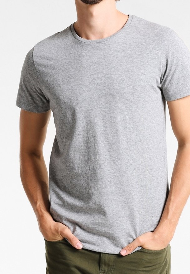 Homme portant un t-shirt uni gris clair à manches courtes et un pantalon vert olive, debout les mains dans les poches sur un fond blanc.