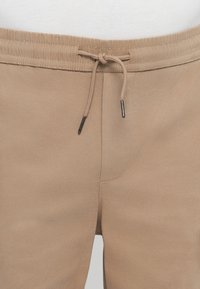 Les pantalons de jogging beige clair présentent une taille élastique avec un cordon de serrage, des embouts en métal et une texture lisse et douce.