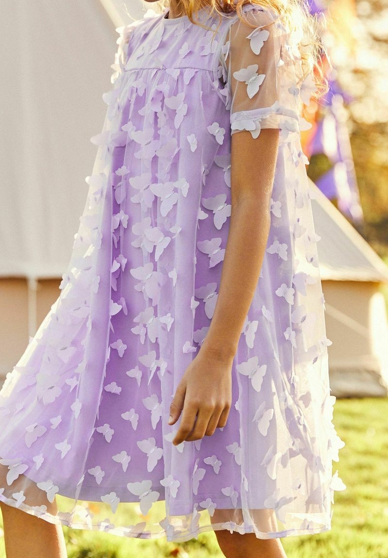 Enfant portant une robe violette claire ornée d'appliques papillons blanches en 3D, debout à l'extérieur près d'une tente sur l'herbe.