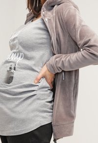 Sudadera con cremallera de felpa gris de textura suave, combinada con una camiseta gráfica de color gris claro. Pantalones negros visibles en la parte inferior.
