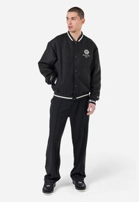 Sort varsity-jakke med hvide striber, knaplukning og broderet logo. Kombineret med sorte bukser og sorte sneakers. Glat tekstur.