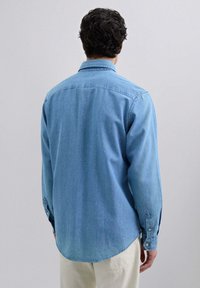Camisa de mezclilla azul claro con mangas largas, un cuello puntiagudo y puños abotonados. Dobladillo curvado y textura suave. Diseño minimalista sin patrones.