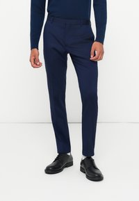 Calças de fato azul-marinho com uma textura suave, design slim fit, passantes para cinto e uma pregueada na frente, combinadas com sapatos pretos.