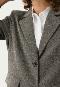 Femme portant un blazer en tweed gris sur une chemise blanche boutonnée, avec un bouton noir fermé à l'avant du blazer.