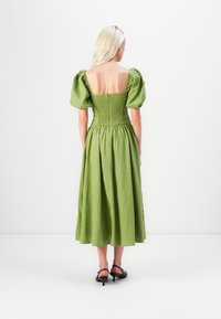 Robe en lin verte avec de courtes manches bouffantes, un dos smocké et une jupe longue qui tombe en dessous des genoux. Portée avec des sandales à talons noires.