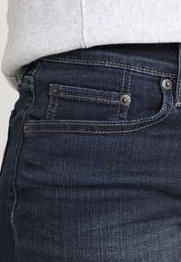 Donkerblauwe spijkerbroek met zichtbare stiksels, vijf zakken en metalen knoopdetails. Zacht grijs stof van de trui dat over de tailleband valt.