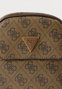 Braune Handtasche mit Muster und einem dreieckigen Guess-Logo in Gold. Die Oberfläche ist glatt mit einem dezent geprägten Design.