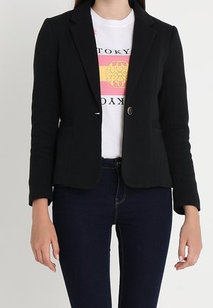 Vrouw draagt een zwarte blazer met een enkele knoop over een wit grafisch T-shirt en donkerblauwe spijkerbroek, staand tegen een effen lichte achtergrond.