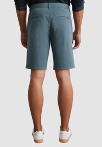 Shorts pour homme en vert foncé, avec une taille plate et des poches arrière, fabriqués en coton. Design décontracté, longueur au genou, associés à des baskets blanches.