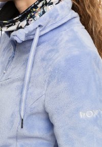 Vaaleansininen fleece-takki, joka on pehmeä ja miellyttävä käyttää. Takissa on huppu, nauhakuja ja kukkakuosilla vuorattu kaulus. Hihassa on brodeerattu logo.