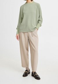 Maglione a maniche lunghe verde chiaro, vestibilità comoda, texture stratificata; abbinato a pantaloni beige a gamba larga e mocassini neri con accenti dorati.