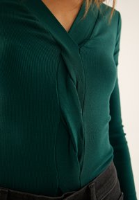 Green ribbed long-sleeve top featuring a twisted design detail at the neckline and a fitted silhouette, paired with dark pants.