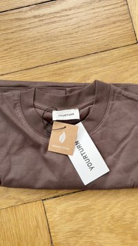 Camiseta de algodón orgánico marrón, doblada, con cuello redondo, que presenta una etiqueta que dice "YOURTURN" y una etiqueta etiquetada como "ALGODÓN ORGÁNICO."