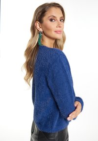 Blauer, flauschiger Pullover mit lockerer Passform, gerippten Bündchen und weicher Textur. Getragen mit langen, grünen, mit Perlen besetzten Ohrringen, die einen markanten Kontrast bilden.