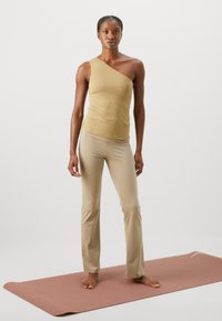 Top beige a spalla unica abbinato a pantaloni attillati beige chiaro. La modella è in piedi su un tappetino da yoga rosa, mostrando una consistenza del tessuto liscia e morbida.