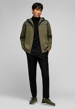 Veste de sport vert olive avec des accents noirs aux épaules, col roulé noir, pantalon noir et baskets vertes avec semelles blanches.