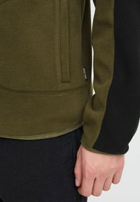 Mørkegrønn fleecejakke med svarte detaljer, som har en glidelås lomme, strukturert materiale og en tettsittende design.