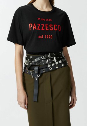 Vrouw draagt een zwart t-shirt met rode tekst en meerdere zwarte leren riemen met zilveren gespen over een olijfgroene rok.