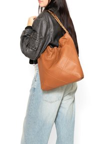 Borsa a tracolla in pelle marrone trapuntata con design morbido e tracolla a catena, indossata da una persona che indossa una giacca nera e jeans blu chiaro.