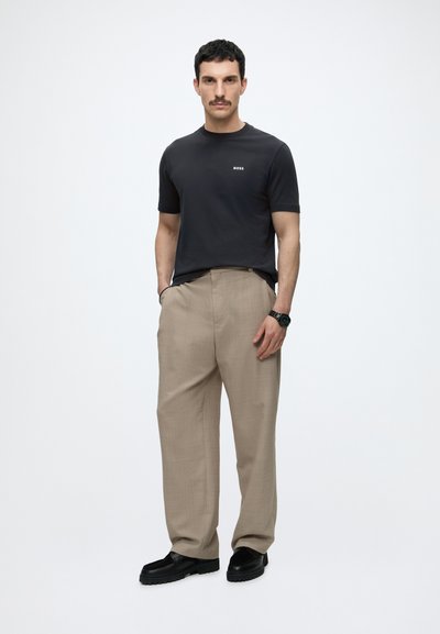 Homme avec une moustache portant un t-shirt noir BOSS, un pantalon beige à jambes larges, une montre noire, des chaussures noires, debout les mains dans les poches sur fond blanc.