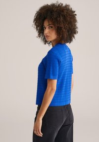 Haut en tricot texturé bleu avec des manches courtes, design cropped et bords côtelés, associé à un jean noir. Présente un motif géométrique.