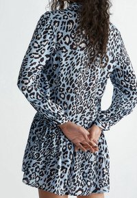 Donna con lunghi capelli ricci che indossa un vestito a maniche lunghe con stampa leopardata blu e nera, mani unite dietro la schiena.