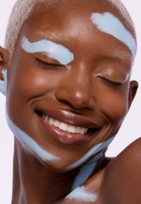 Modell med kort, lys blondt hår, som smiler. Blå ansiktsmaling aksenter på pannen, øyenbrynene og nakken. Glatt hudtekstur, varme undertoner.