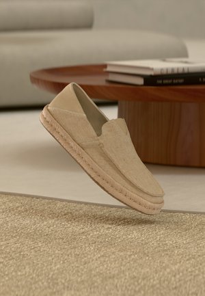 Chaussure beige tissée à enfiler avec semelle tressée flottant au-dessus d'un tapis texturé, d'une table ronde en bois et de livres empilés dans une pièce aux tons neutres.