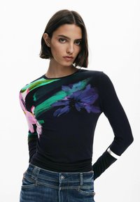 Cosi haut à manches longues noir avec un imprimé floral coloré sur une épaule et des rayures vertes. Le matériau semble doux, avec une coupe ajustée. Jean en denim inclus.