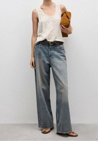 Kobieta w białej, haftowanej bluzce bez rękawów, szerokich, wyblakłych niebieskich dżinsach, z czarnym paskiem, brązowych sandałach i trzymająca brązową torebkę kopertową.