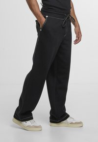 Pantalones de chándal negros con detalles de costura blanca, cintura elástica, bolsillos laterales y un corte holgado. Combinados con zapatillas crema y grises.