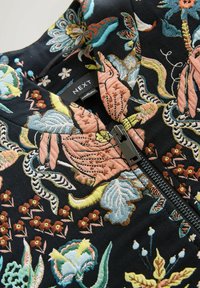 Veste en tissu noir avec des broderies florales colorées, dotée d'une fermeture éclair et d'une texture lisse. Étiquette visible : "NEXT."