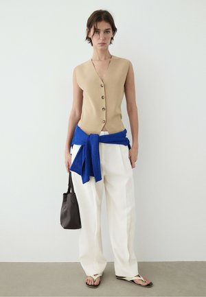 Donna che indossa un top beige senza maniche con bottoni, pantaloni bianchi larghi, maglione blu legato in vita, tiene una borsa nera e indossa sandali infradito.