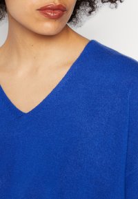 Femme aux cheveux bouclés portant un pull bleu vif à col en V, avec le cou, les lèvres et une partie de la mâchoire visibles.