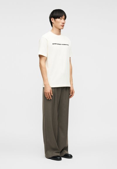 Άνδρας στέκεται φορώντας λευκό T-shirt Emporio Armani, γκρι παντελόνι με φαρδύ πόδι και μαύρα παπούτσια, μπροστά σε απλό φωτεινό φόντο.