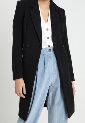 Frau trägt einen schwarzen langen Mantel über einem weißen Hemd mit Knöpfen und hellblauen Cordhosen.