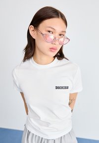 Balta medvilninė t-shirt marškinėliai trumpomis rankovėmis, su juodu logotipu "DICKIES" kairėje krūtinės pusėje. Šviesiai rožiniai katės akies akiniai.