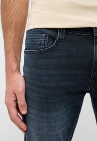 Jeans azul oscuro con una textura suave, que presentan un bolsillo trasero y sutiles detalles de costura. Combinados con una camisa ajustada beige.