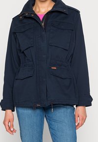 Granatowa kurtka robocza z zamkiem z przodu, dwoma kieszeniami na piersi i dwiema kieszeniami w talii. Posiada ściągacze w talii i przy mankietach.